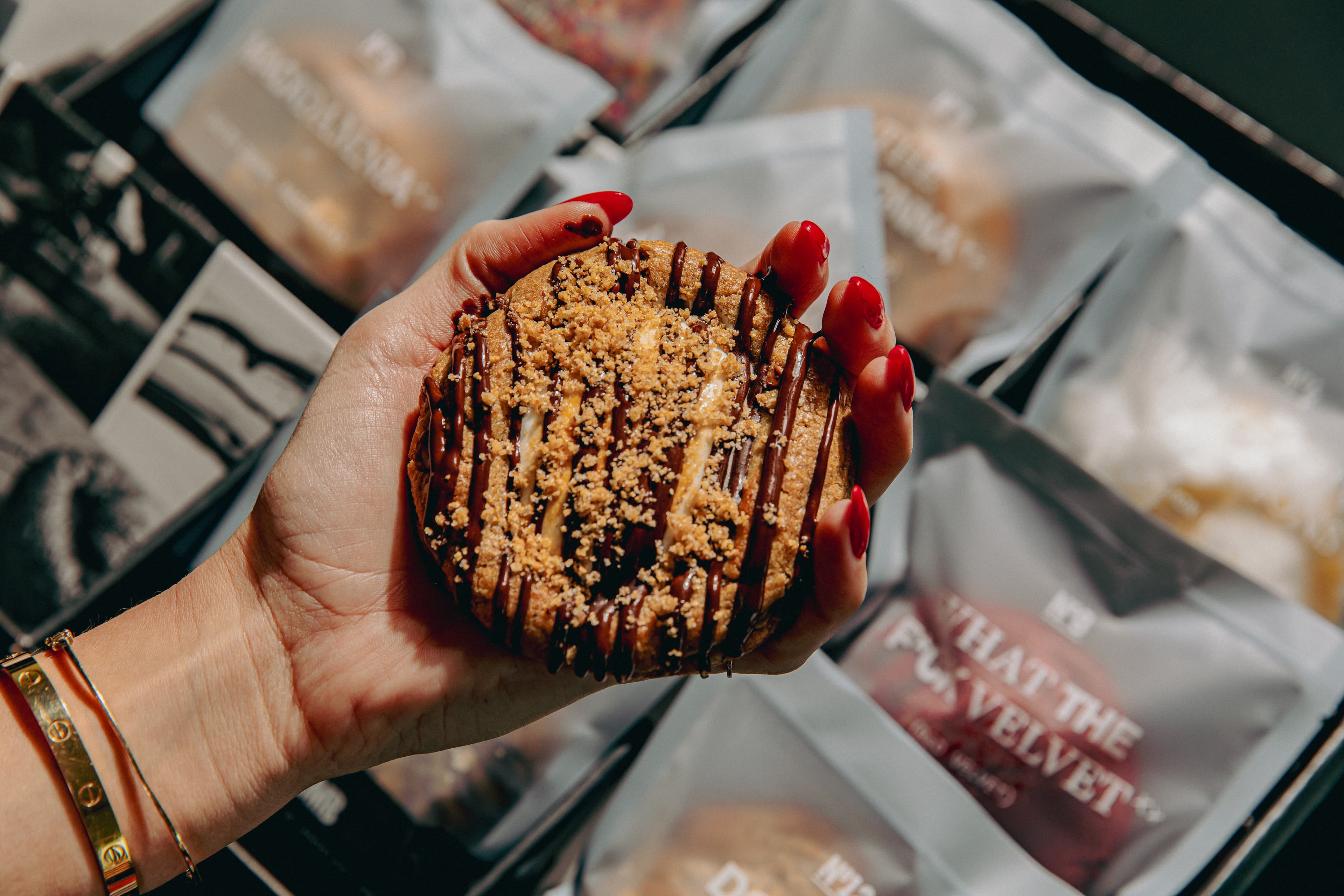 A hand holding a chocolate-drizzled s’mores cookie over a box of wrapped Last Crumb cookies.